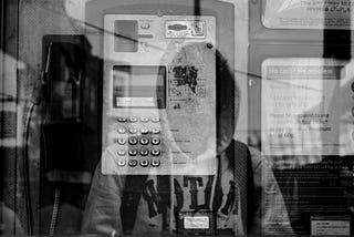 A grayscale photograph with overlay effects showing a reflection of a battered pay phone overlaying a caller in a hoodie (with their face obscured). To the right is writing from a pay phone. You can also just barely glimpse reflections of a building.