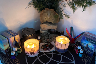 Altar cloth, black with the triquetra symbol partially showing in silver and gold. Two large lit candles, sets of oracle cards, essential oils, crystals, and colored pens. The back of the altar includes items from nature,:an interesting slab of wood, a stone, foliage, and moss collected from nearby.