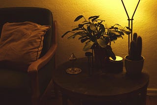 An arm chair and small desk with plants and a candle, dimly lit by a lamp.