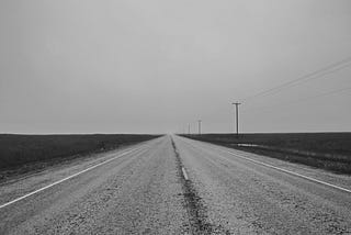 Road, surrounded by a flat terrain, heading off into the distance. Black and white photo.