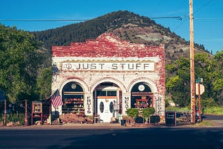 Old brick store painted red in parts with three arches around door and windows that says Just Stuff against green hill and with American flag out front