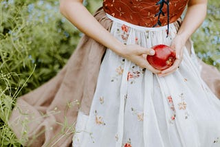 Half-frame view of a seated person in a floral dress, gently holding a bright red apple with both hands, surrounded by lush green foliage in the soft daylight, evoking a pastoral and serene atmosphere.