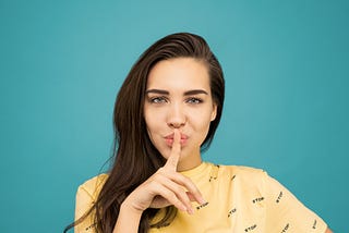 Portrait photo of a woman in a yellow t-shirt doing the shh sign while standing in front of a blue background.
