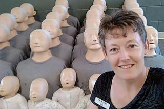The author, Gill McCulloch, wearing a dark t-shirt and smiling. In the background are lines of CPR manikins.
