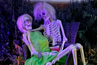 Two skeletons very much in love, the female is sitting on the male’s lap, in the dark with neon light in halloween fashion.