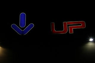 On a black background, a big blue arrow on the left points straight down while on the right half of the field is the world “UP” in red capital letters.