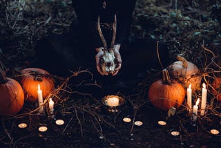 A woman kneeling on the ground at night holds a goat skull while surrounded by pumpkins and candles