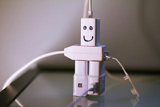 Three white plugs plugged into a white extension cord with a face drawn on so it looks like a robot