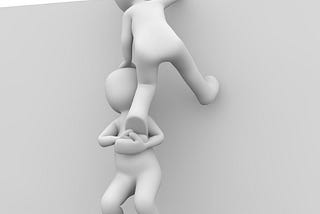 A black and white computer generated image of two faceless humanoid figures against a blank wall. One is boosting the other up and over the wall.