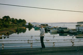 A middle-aged white man in glasses, a light white jacket and black pants stands on a bridge overlooking a small marina