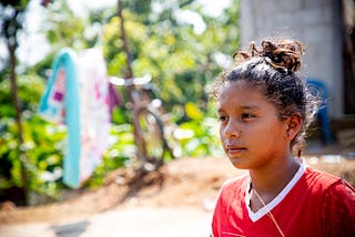 Daniela is wearing a red shirt and jeans. She is standing outside her house and is looking into the distance.