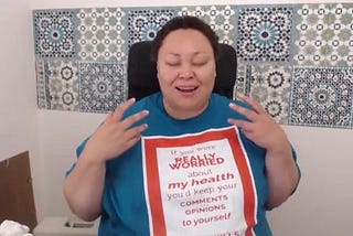 A photo of a fat woman of color laughing with her eyes closed while sitting in a chair.
