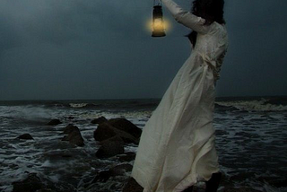 Dark-haired woman in a long white dress, standing on rocks and holding a lantern toward a dark sea.