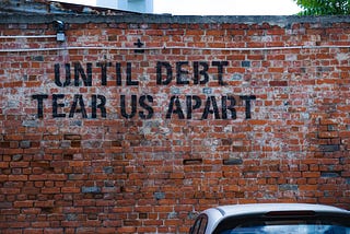 Red brick wall with a car parked in front of it. Text on the brick wall is black stencil grafitti that reads, “Until debt tear us apart” in all caps.