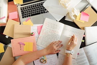 A woman’s arms are visible as she writes in a notebook. She is surrounded by papers, notebooks, and a laptop. She is wearing a white watch.