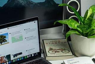 A laptop, startup book and notebook is kept on the table. Desktop and speaker is in the background. A cute table plant is on the side of the book.