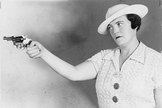 Black and white photo of a woman in her forties with a tough expression on her face, wearing a white dress and hat, and holding a gun in her hand