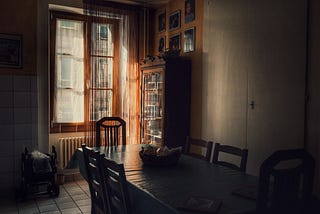 Soft morning light filters through sheer curtains onto a quiet dining room. A long table with empty chairs sits in shadow, except for one gently lit by the window. A basket rests at the center, and a folded stroller sits off to the side. Photos line the far wall. The room feels recently occupied, but now still.