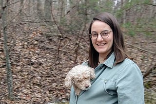 The author, a white woman in her mid-twenties, smiling in the woods while wearing a short green jacket that is straining at the seams due to a small beige poodle being stuffed inside. The poodle’s head sticks out above the top button like an indifferent teddy bear.