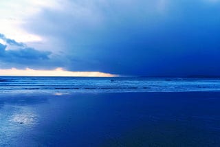 A broad royal blue seashore landscape with a sharp stroke of white sky cracking through the horizon like the deft brush stroke of an artist. There are no people to be seen, only the expance of coastal emptiness as the distant waves rolls in with white foamy caps.