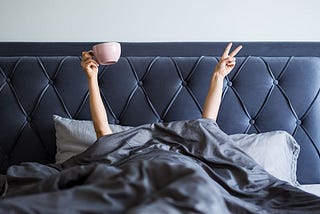 Woman in a large grey bed, head covered with a grey blanket- only her arms show- they are raised up- one with a large pink coffee cup and the other raising the peace sign