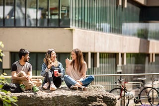 Wide front on view of three students sat on a rock outside a modern building which is blurred in the background. All three students are sat cross legged and the middle student is holding a red coffee cup. There are a couple of bikes leaning against railings to the right background. It is a bright and sunny day.