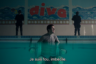 La caméra est plongée dans une piscine, de sorte qu’on voit en partie sous l’eau et en partie au dessus, et notamment le mur du fond. On voit donc que David est attaché sur une chaise dans la piscine. Dans le fond, sur le rebord, deux flics, type CRS vénères qui surveille. Sur le mur du fond, une mosaïque où est écrit “dive” (“plongez”). David parle à quelqu’un hors champs pour dire “je suis fou, imbécile”