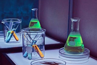 3 test tubes in a beaker next to a large tube of green liquid on a petri dish