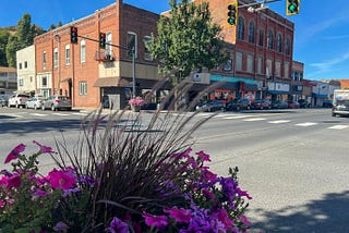 A colorful mix of flowers sit in the foreground in front of a traffic intersection and a row of historic buildings along Main Street in Colfax, Wash. Cars line the front and side of the buildings. Traffic signals are visible at the top of the frame above the intersection.