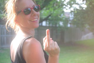 Young woman smiling, wearing red sunglasses and flipping up her middle finger.