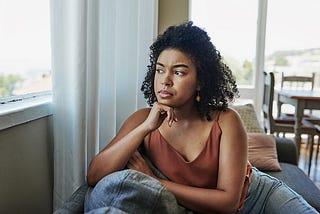 Black woman in brownish gold strappy top, staring out of window with her chin resting on her fist.