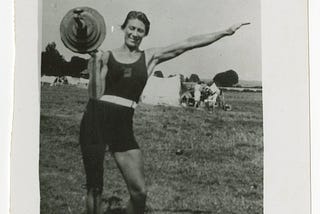 Black-and-white photo of Ivy Russell standing outside and facing the camera. She is holding a loaded barbell on her right shoulder, and her left arm is stretched out. She is wearing a dark swimsuit-like training costume and white shoes. She has short dark hair and is smiling.