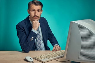 A man in a tie and suit with a moustache, looking at the camera. He is sitting behind an old fashioned looking computer, keyboard and mouse.