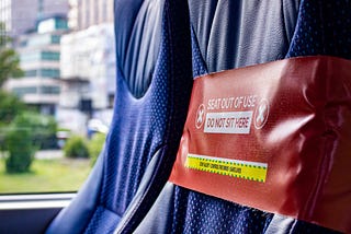 This is a close up photo of two seats by a window in a train. The baground is out of focus. The closest seat from the objective has a thick red ribbon on it, with “Seat out of use, do not seat here” and the UK’s COVID yellow and green tagline “Stay alert, control the virus, save lives”.