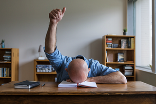 Bald man with face down on desk raising a thumbs up gesture — My Amazon Novels Suck