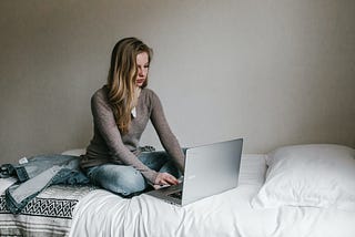 Woman typing on her laptop while sitting on her bed.