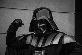 A person dressed as Darth Vader in full black armor and helmet, raising a fist in a dramatic pose.