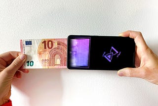 a hand pushes a 10euro note into a smartphone from left to right. on the screen of the smartphone is the end of the euro note in purple pixels — the fiat money is transformed into pixels. on the right side of the screen on a black background a digital hourglass is showing that this is being processed.