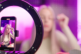 A blonde woman applying mascara in front of a ring light and tripod recording a video stream of her tutorial.