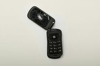 Black flip phone with smashed screen and top half broken away from bottom half, against a white background.