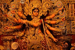 A photograph of the goddess Durga from Hindu mythology. She has eight arms, each holding a weapon, namely, sword, shield, bow & arrow, conch, discus, bell, and mace. The other two arms hold the trident, impaling the demon Mahishasura at her feet with a serene expression while sitting on a lion. The large idol is sculpted beautifully and adorned with jewels and detailed clothing. This style of idol sculpting is usually seen in West Bengal, India.