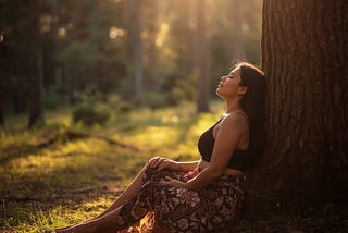 Woman sitting quietly in a sunlit forest surrounded by soft golden light, reflecting peacefully in nature and symbolizing anxiety, mindfulness, nervous system healing, and returning to the present moment.