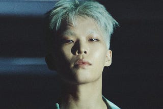 A profile shot of Lee staring into the camera, his hair bleached white and illuminated against a dark background.