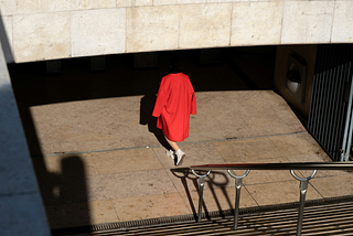 A photo of the back of a woman at the bottom of a set of stairs leading into a metro station. She’s wearing bright red overcoat and white Converse shoes with bare legs. The shadow from the entrance of the station makes it look like the woman has no head.