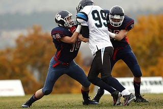 Three football players during a running play, two in dark blue uniforms and one in a white uniform.