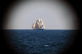 a view of a large sailboat in the distance with billowing sails on the open ocean. the sails are beige. the water is a shade of deep blue. the sky appears flat and grey. the image appears similar to how it might appear through a telescope. there is a circular vignette effect on the image.