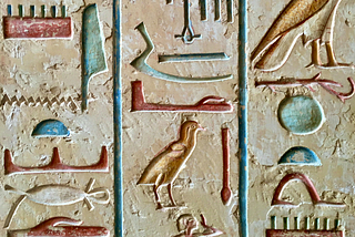 A colourful mix of close-up Egyptian hieroglyphs carved into sand-coloured stone. The hieroglyphs are separated into three columns, with a blue carved line between them.