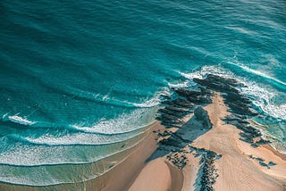 View from a high cliff, with a spit of sandy beach extending out into a rippling turquoise sea.