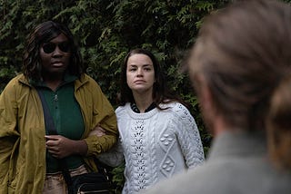 Still image of the lead actresses during production. We see a Black female with dark shades, wearing a mustard color jacket and pine green shirt. Next to her, holding her arm, is a Middle Eastern woman, wearing a white sweater. Directly infront the camera is a White male facing the woman, his back towards us. We see the back of his dark brown hair and gray color of his shirt.