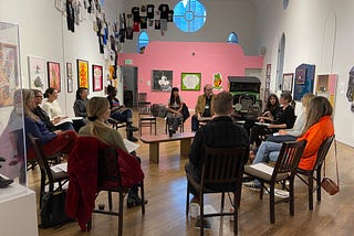 A group of people seated in a circle of chairs talks in an art gallery filled with art on the walls.